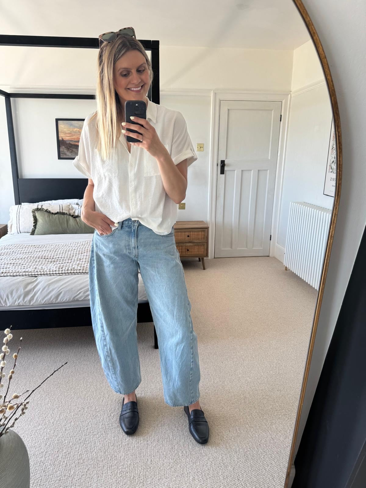 Person taking a mirror selfie wearing a white blouse, light blue jeans, and black shoes in a bedroom wearing large sized womens shoes
