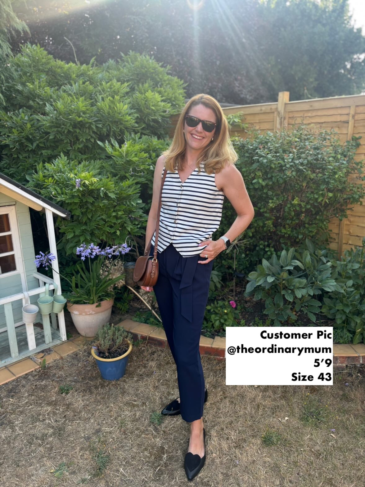Woman with size 9 shoes standing outdoors in a garden, wearing a striped top and navy pants.