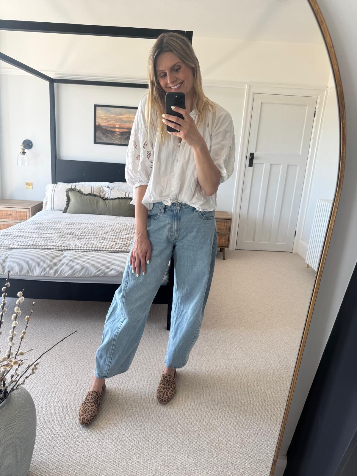 Woman taking a mirror selfie in a bedroom wearing a white blouse and blue jeans wearing large sized womens shoes