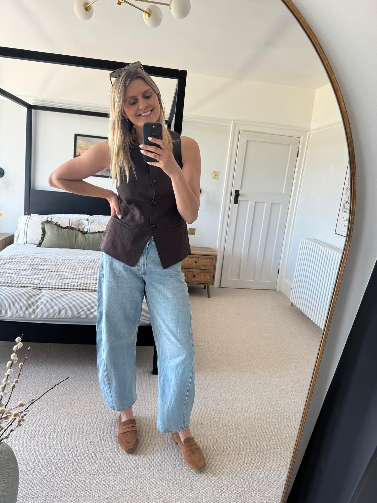 Woman taking a mirror selfie in a bedroom wearing a brown vest and light blue jeans and large sized womens tan suede shoes